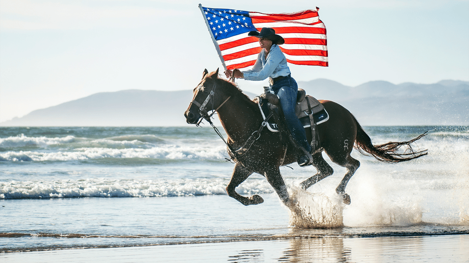  A cowboy rides a horse on a beach, carrying an American flag and listening to royalty free patriotic music.