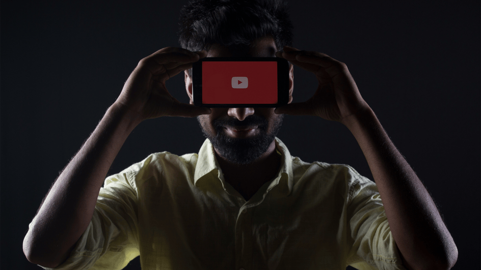  A man holds a black smartphone displaying the YouTube app on the screen, as he learns about top faceless YouTube niches and faceless YouTube channel ideas